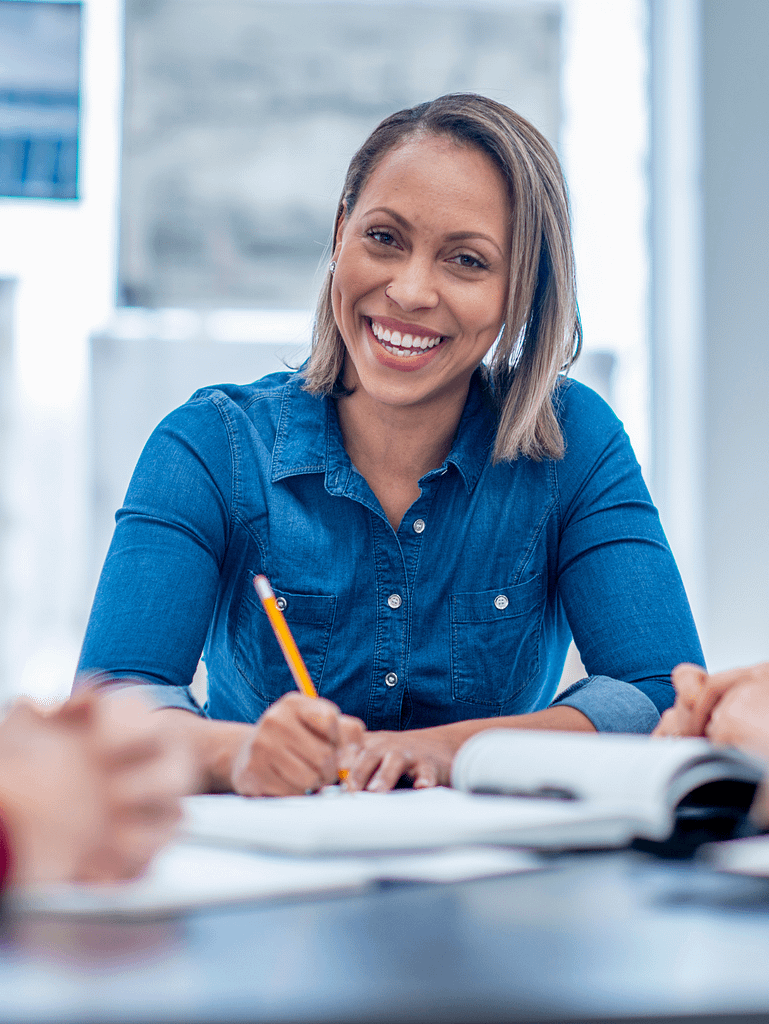 A smiling confident woman in a blue shirt sitting at a desk, writing in a notebook on a meeting. Looking straight into the camera