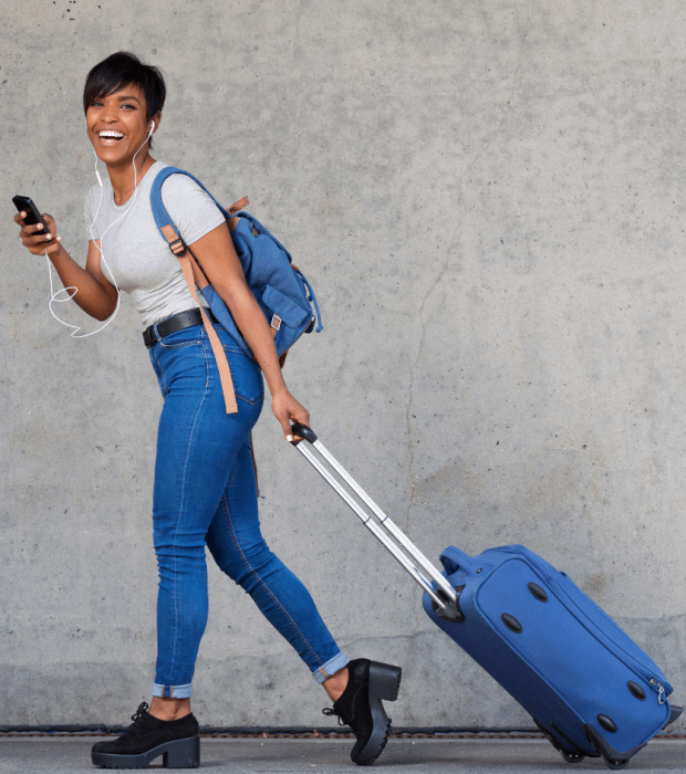 A person walking with a rolling suitcase and wearing a backpack, looking at a smartphone with earphones plugged in, symbolizing the convenience of having travel health insurance.